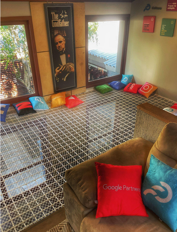 Sala decorada com almofadas coloridas de marcas digitais como Google Partners, Instagram e outras, sobre um piso de rede suspensa, com pôster do filme 'O Poderoso Chefão' na parede.