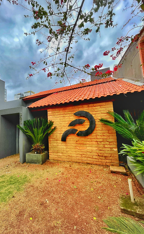 Fachada de prédio com parede de tijolos aparentes, telhado de telhas vermelhas e logo da Fator.ag em preto fixado na parede, cercado por plantas ornamentais.