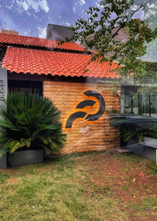 Fachada de prédio com parede de tijolos aparentes e telhado de telhas vermelhas, exibindo o logo da Fator.ag preto em destaque, rodeado por plantas no jardim.