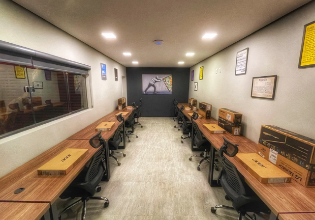 Sala de escritório com mesas alinhadas nas laterais, cadeiras pretas e caixas de notebooks sobre as bancadas, decorada com quadros e iluminação de teto.