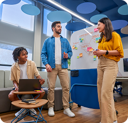 Três estudantes em sala moderna colaboram em frente a um quadro branco com post-its e anotações coloridas, enquanto um deles utiliza um notebook.