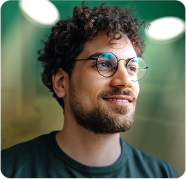 Homem jovem de óculos e barba curta, sorrindo levemente em ambiente iluminado.