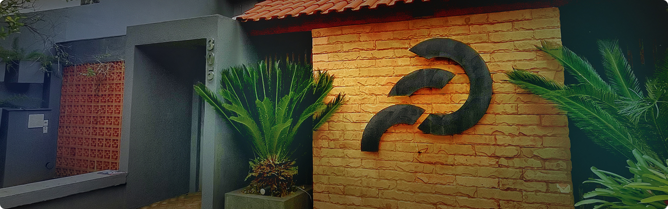 Fachada de prédio com parede de tijolos amarelos, logo escuro em relevo e plantas verdes ao redor.