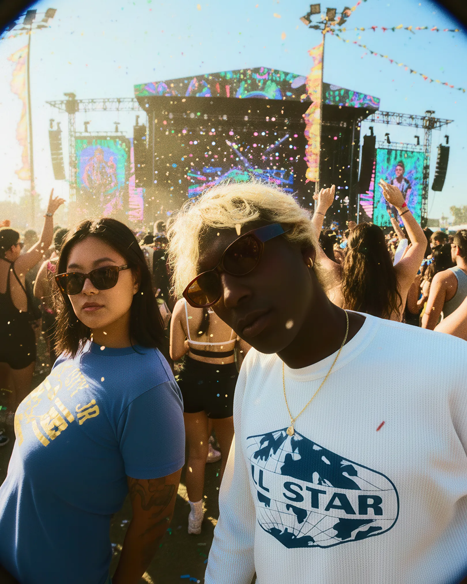Two young people wearing sunglasses at an outdoor music festival with a stage and crowd in the background.