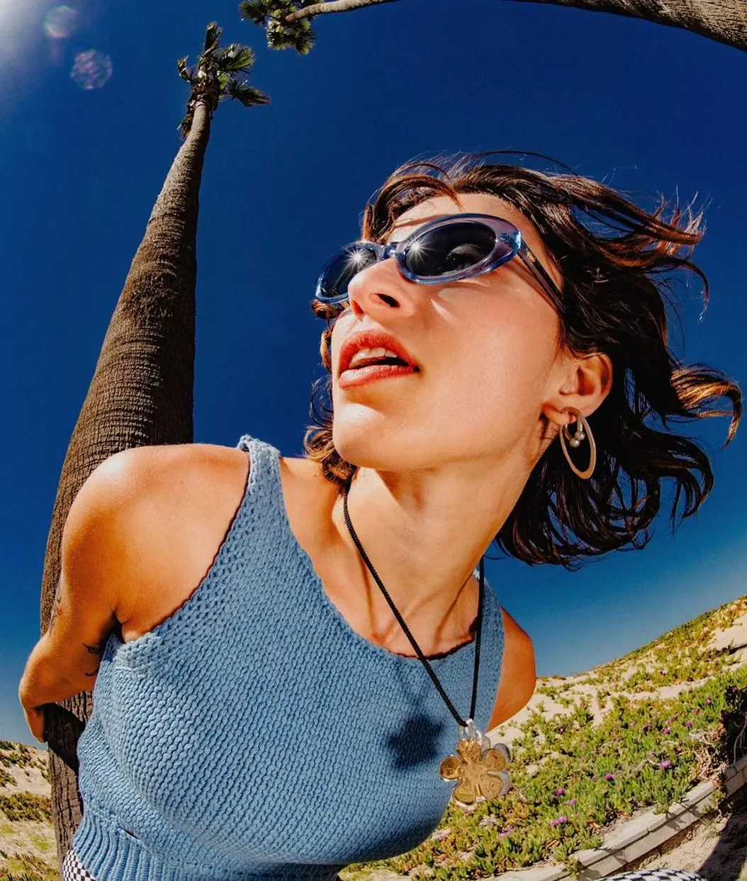 Young woman wearing blue knit tank top and sunglasses leaning forward outdoors with palm trees and blue sky in background.