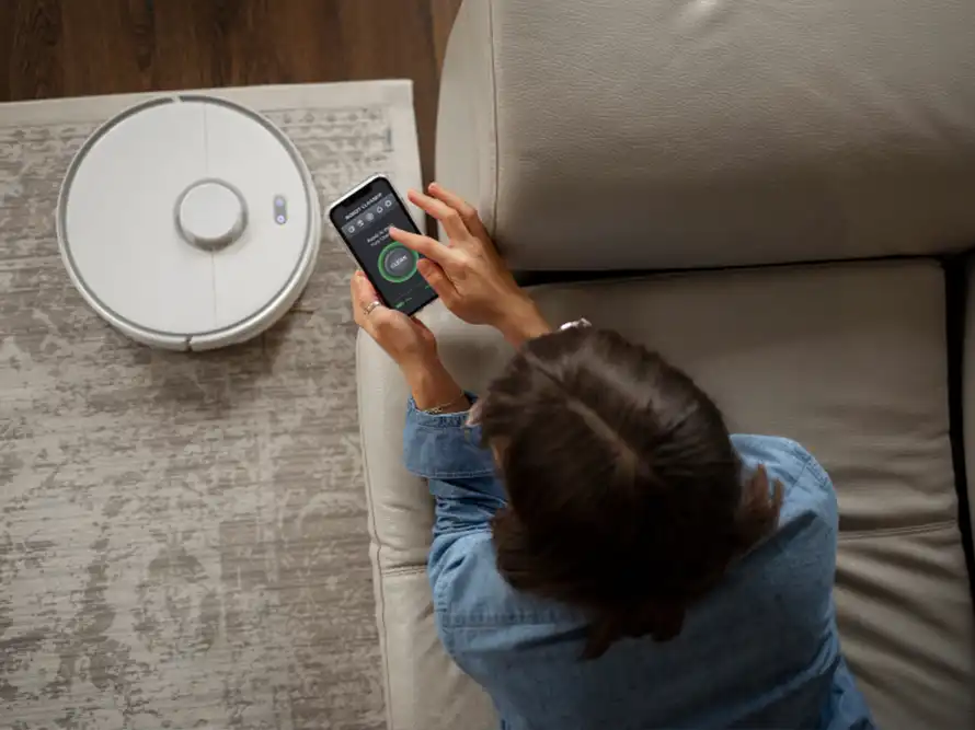 Person sitting on a couch controlling a white robot vacuum cleaner using a smartphone app.