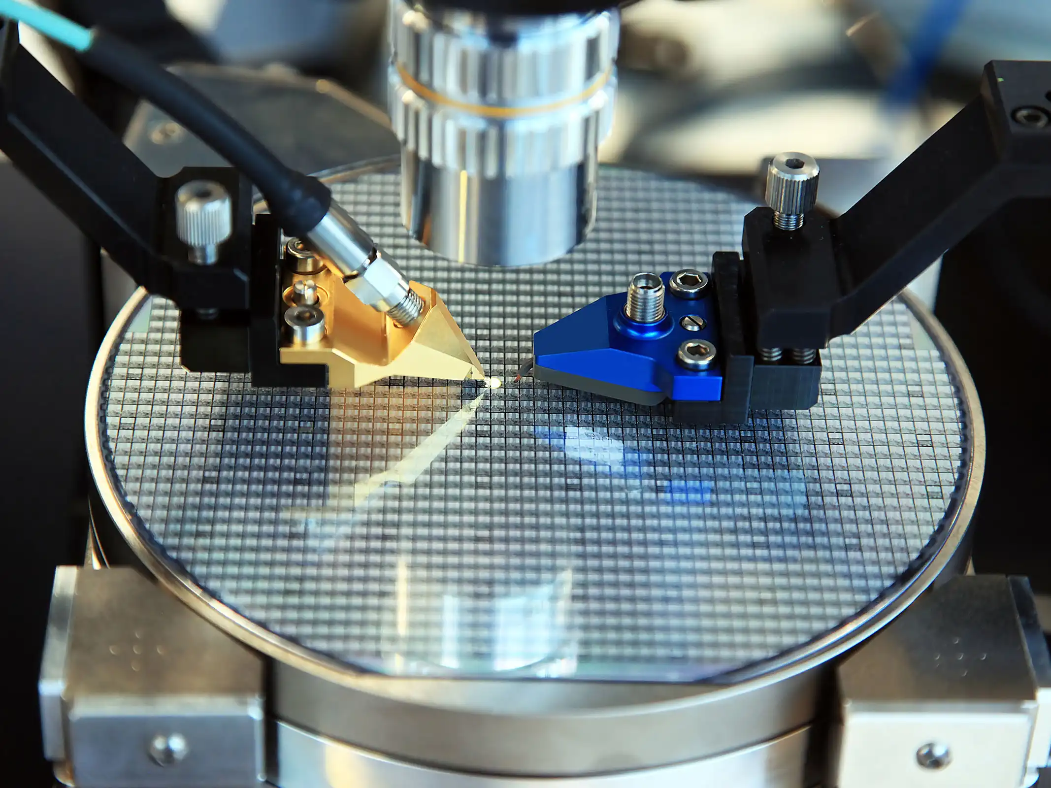 Close-up of a silicon wafer being inspected with precision tools and a microscope lens in a laboratory setting.
