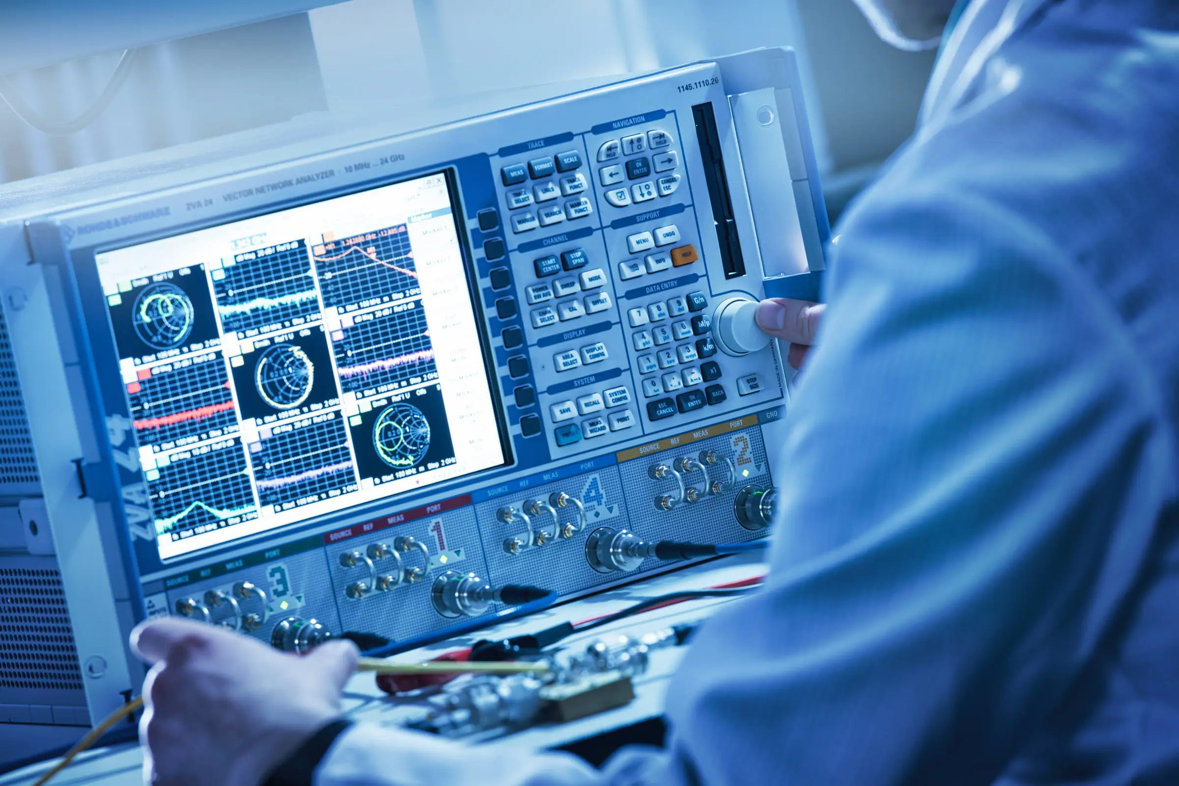 Engineer adjusting controls on a vector network analyzer displaying multiple signal graphs.