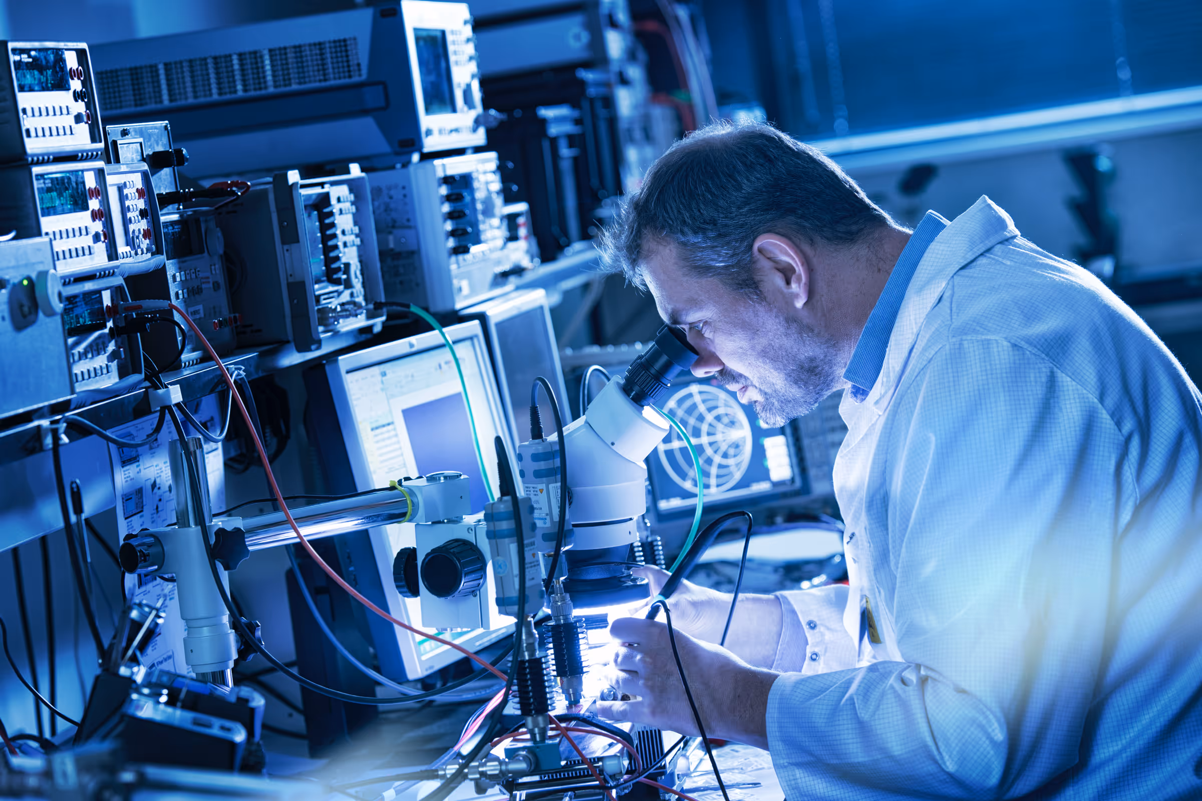Scientist in a lab coat using a microscope amid electronic equipment and monitors in a laboratory setting.