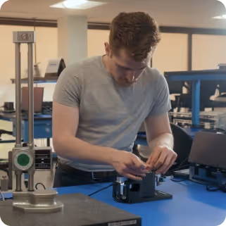 Man in gray t-shirt working with mechanical equipment on a blue workbench in a laboratory or workshop.