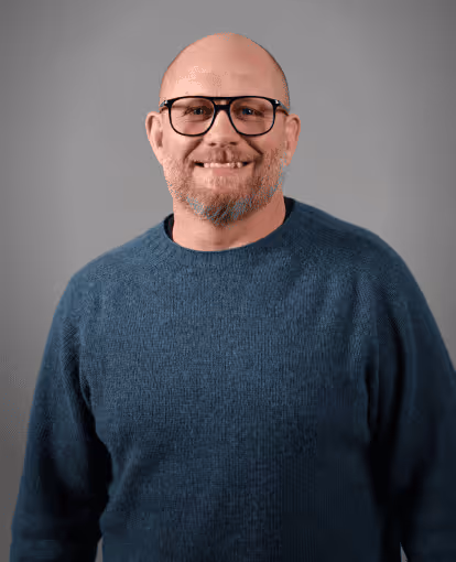 Middle-aged man with glasses, beard, and blue sweater smiling against a gray background.