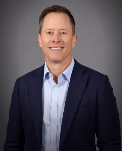 Smiling man wearing a dark blazer and light blue shirt against a gray background.