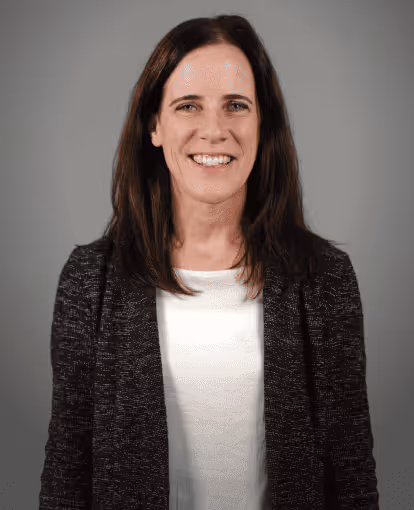 Smiling woman with shoulder-length brown hair wearing a white shirt and dark patterned blazer against a gray background.