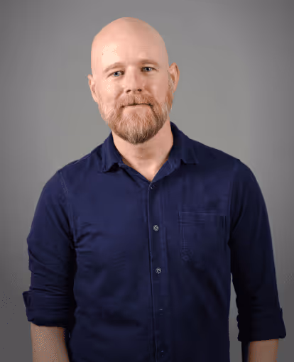 Bald man with a red beard wearing a dark blue button-up shirt standing against a gray background.