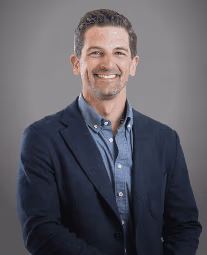 Smiling man with neatly styled hair wearing a dark blazer over a blue button-up shirt, posed against a plain gray background.