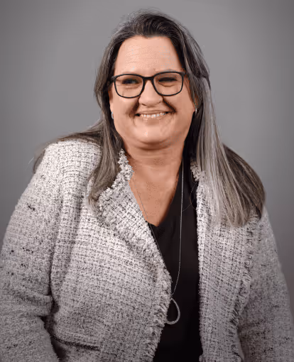 Woman with long gray hair wearing glasses, a black top, and a silver teardrop pendant necklace, smiling softly against a gray background.