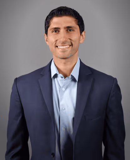 Smiling man with short black hair wearing a navy blazer and light blue dress shirt standing against a gray background.