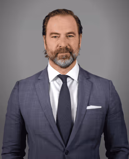 Man with beard and slicked-back hair wearing a blue suit, white shirt, and navy tie against a gray background.