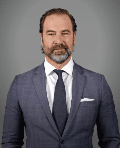 Man with beard and slicked-back hair wearing a blue suit, white shirt, and navy tie against a gray background.