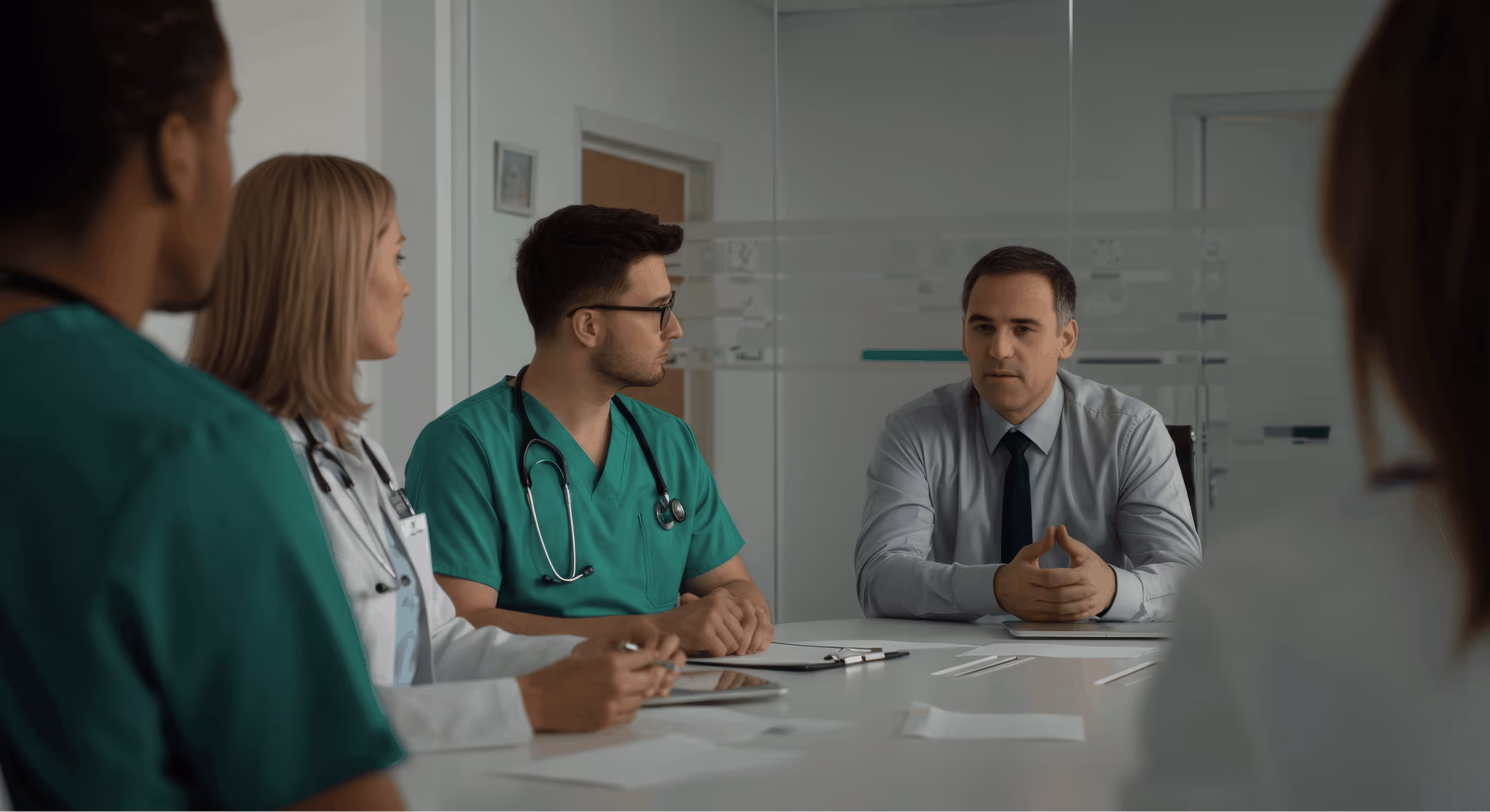 Healthcare professionals and a man in a shirt and tie having a serious discussion around a conference table in a medical office.
