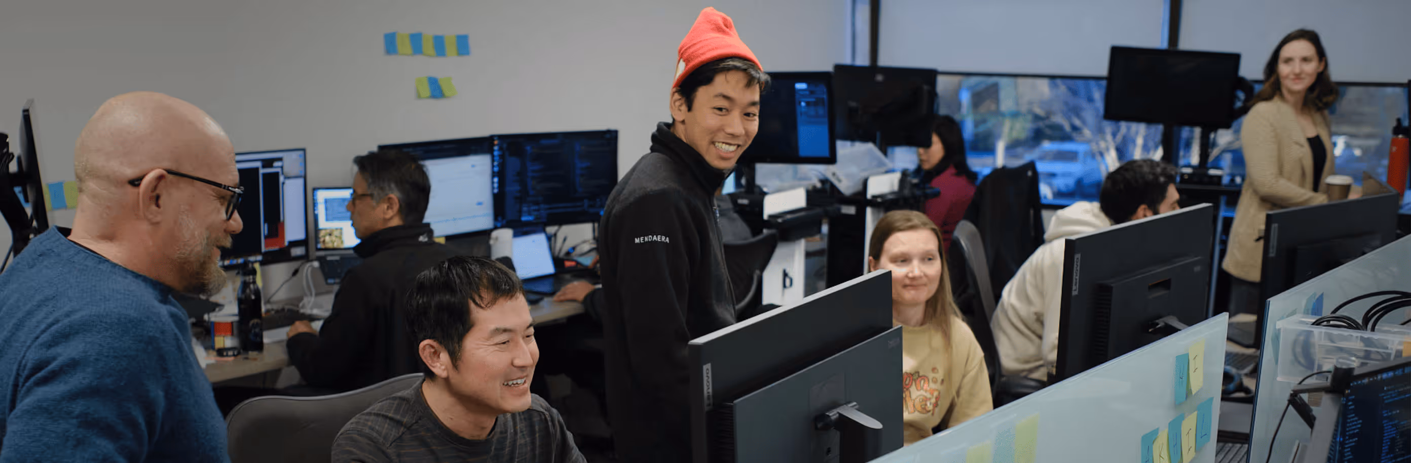A diverse group of people working collaboratively in an office with multiple computer monitors and a casual atmosphere.
