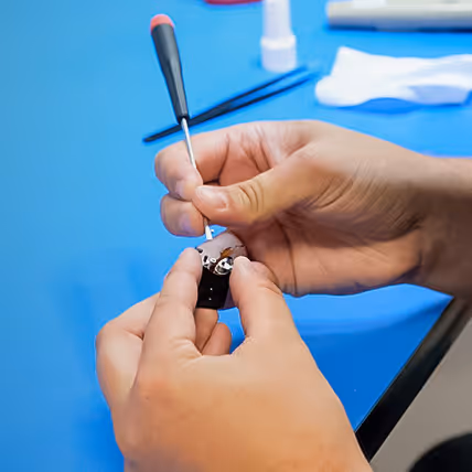 Hands using a small screwdriver to repair a watch movement on a blue surface.