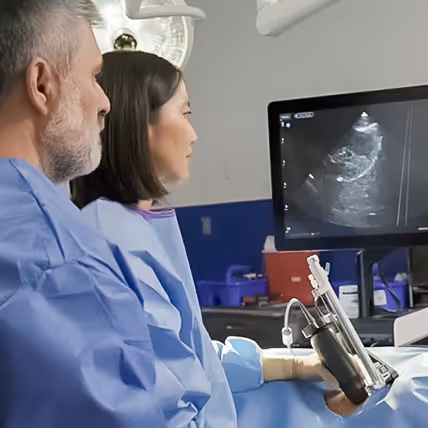 Two medical professionals in blue gowns examining an ultrasound image on a monitor in a clinical setting.