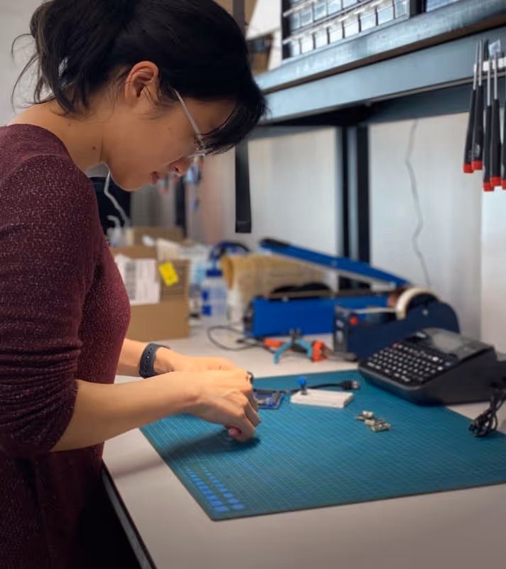 Woman working with electronic components on a blue cutting mat at a workbench in a workshop.