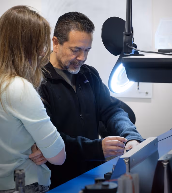 Two people focused on a technical device, one adjusting controls under a bright ring light.