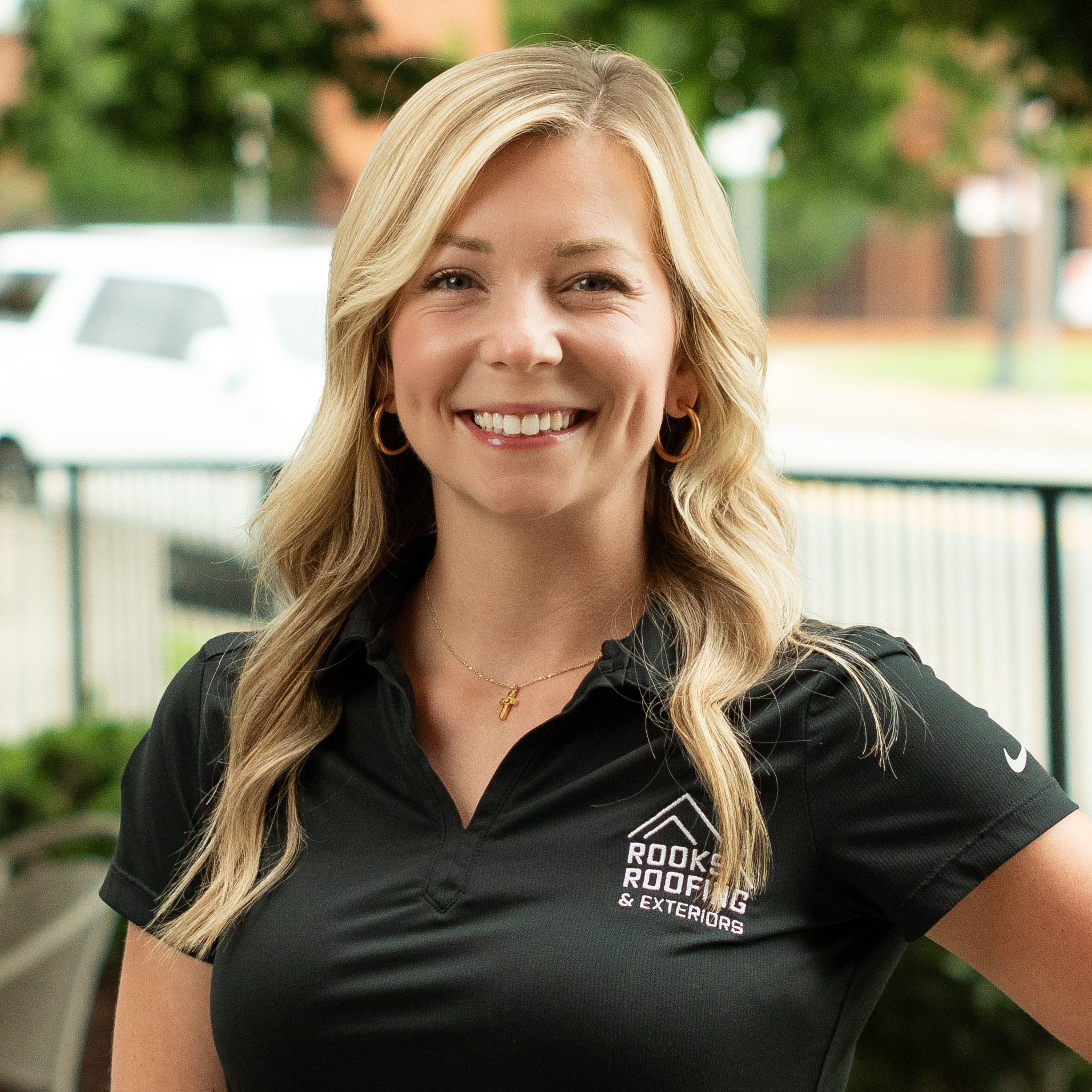 Smiling blonde woman with wavy hair wearing a black Rooks Roofing polo shirt and gold hoop earrings.