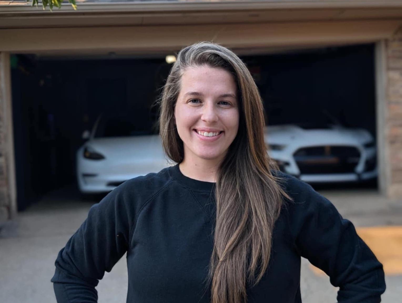 Smiling woman in black sweater standing in front of garage with Tesla cars