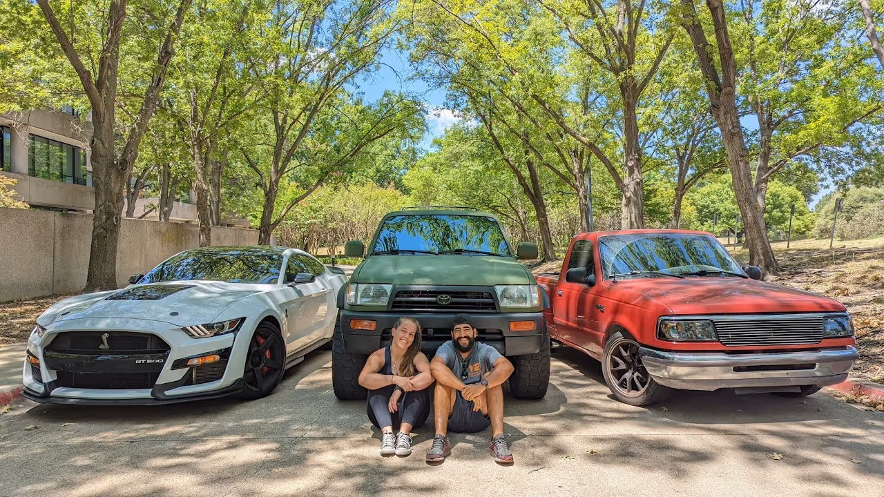 Two people sitting between white, green, and red vehicles under trees