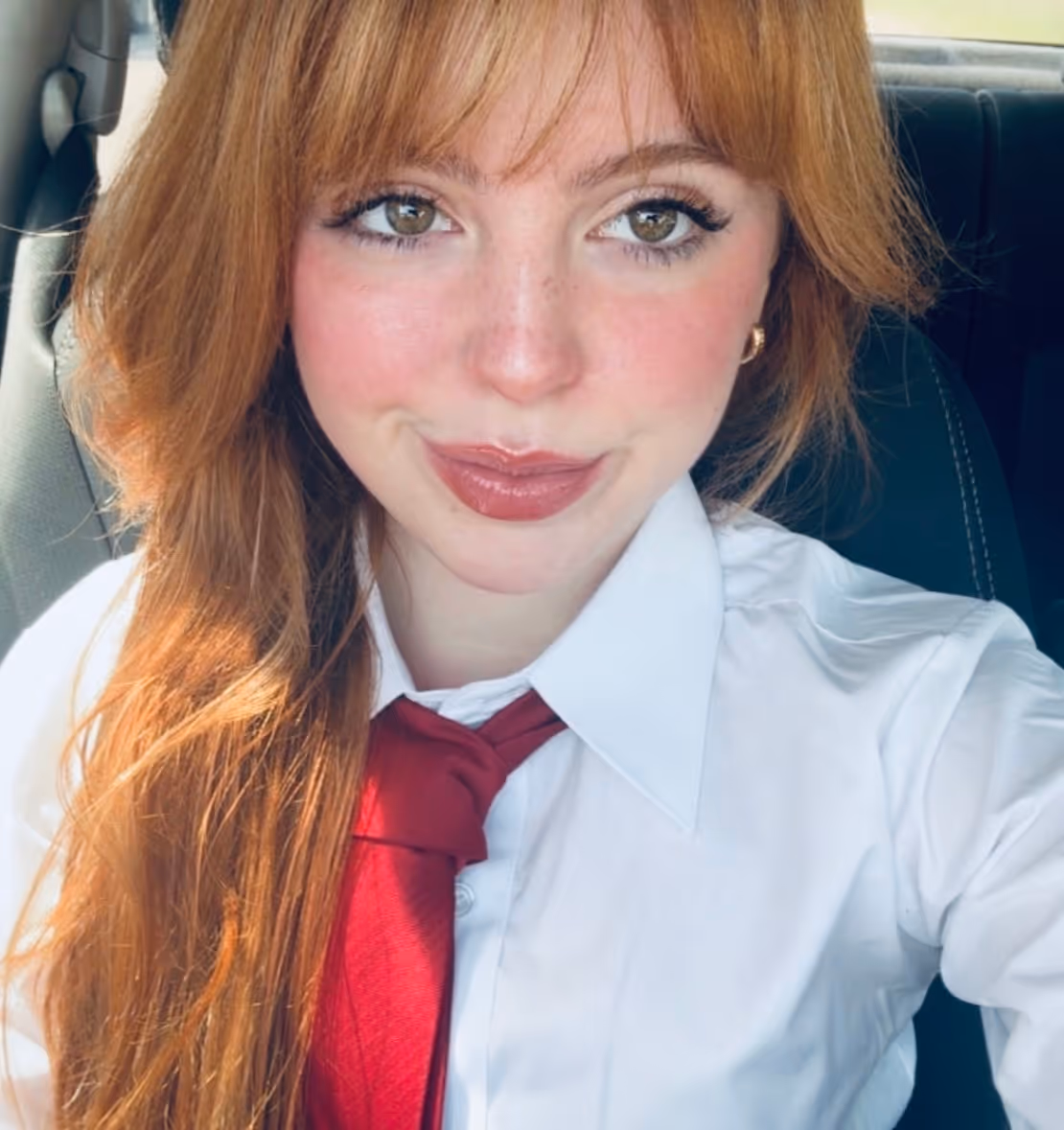 Young woman with long red hair, white shirt, and red tie, smiling inside a car.