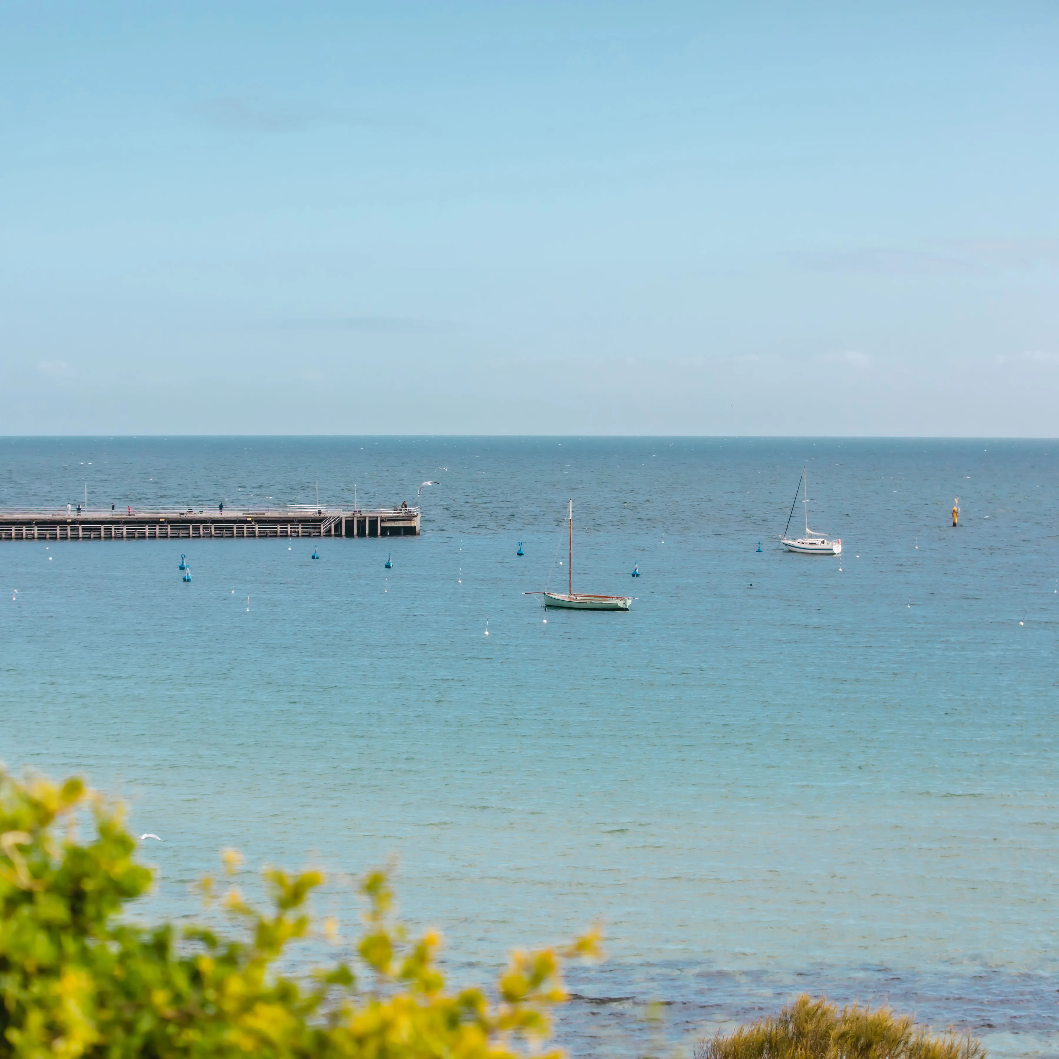 Two sailboats anchored near a wooden pier on calm blue ocean waters under a clear sky.