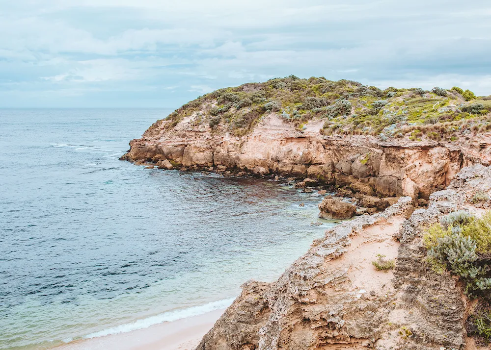 Rocky coastline with green shrubbery overlooking a calm sea under a cloudy sky.