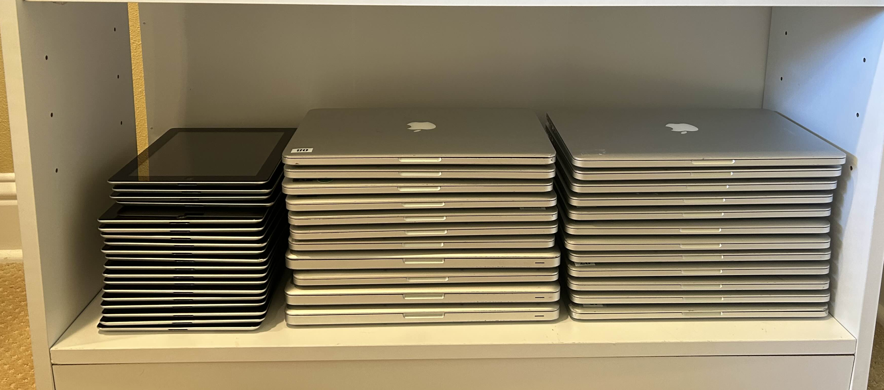 White shelf holding three neat stacks of closed Apple laptops and tablets.