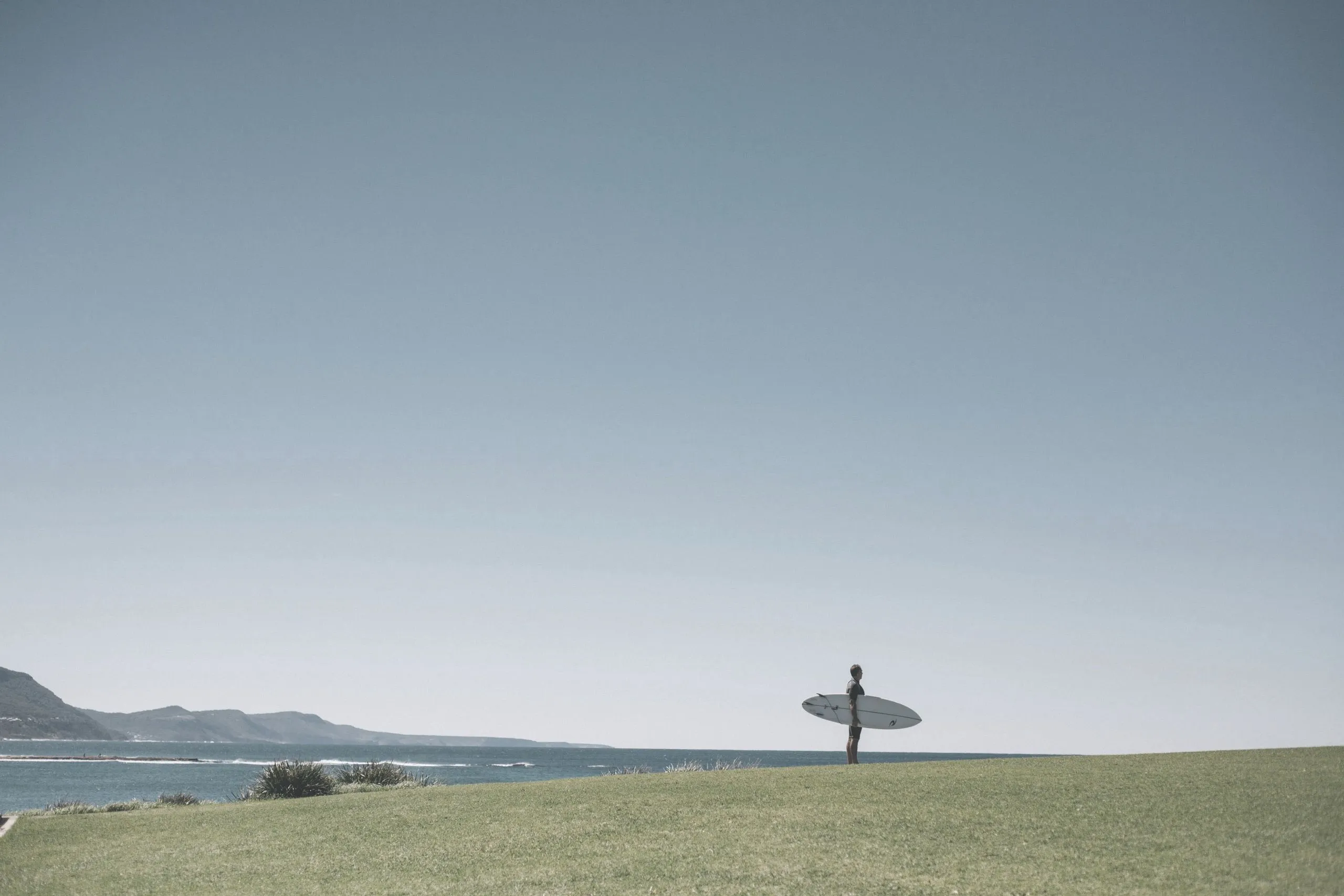 Surfer holding a surfboard stands on green grass overlooking the ocean with distant hills.