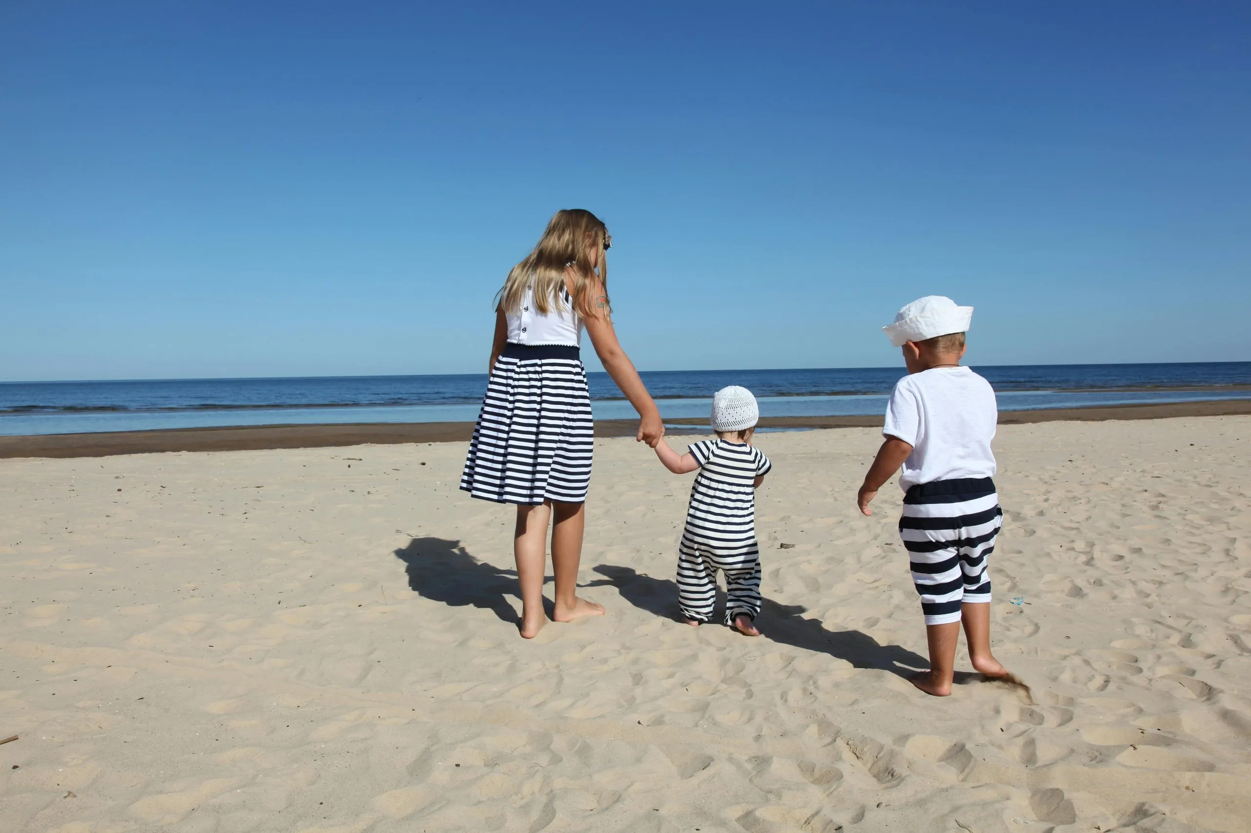 Three children in striped clothing standing barefoot on sandy beach facing the ocean under clear sky.