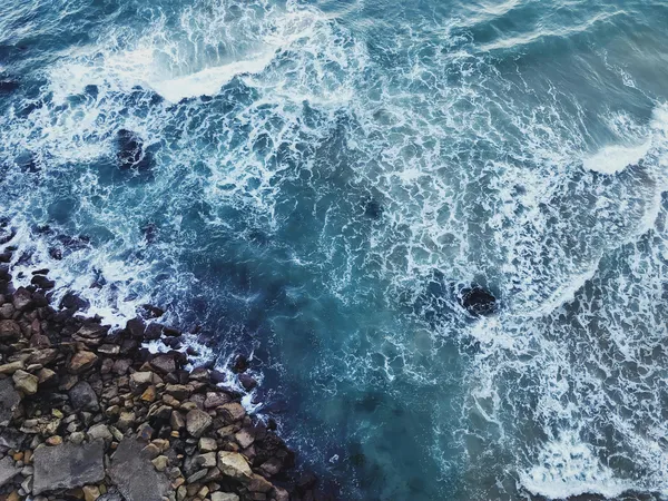 Waves crashing against rocky shoreline with blue turbulent ocean water.