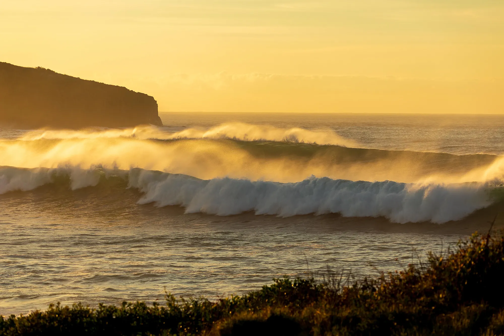Ocean waves rolling toward shore with cliff in distance under golden sunset light.