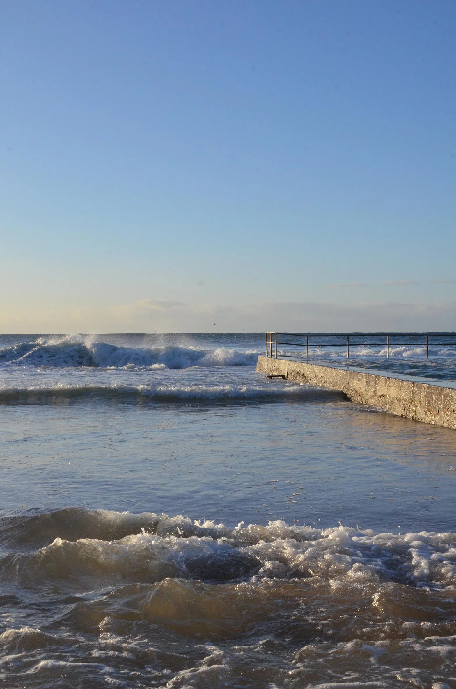 Ocean waves near a concrete pier under a clear blue sky at sunset or sunrise.