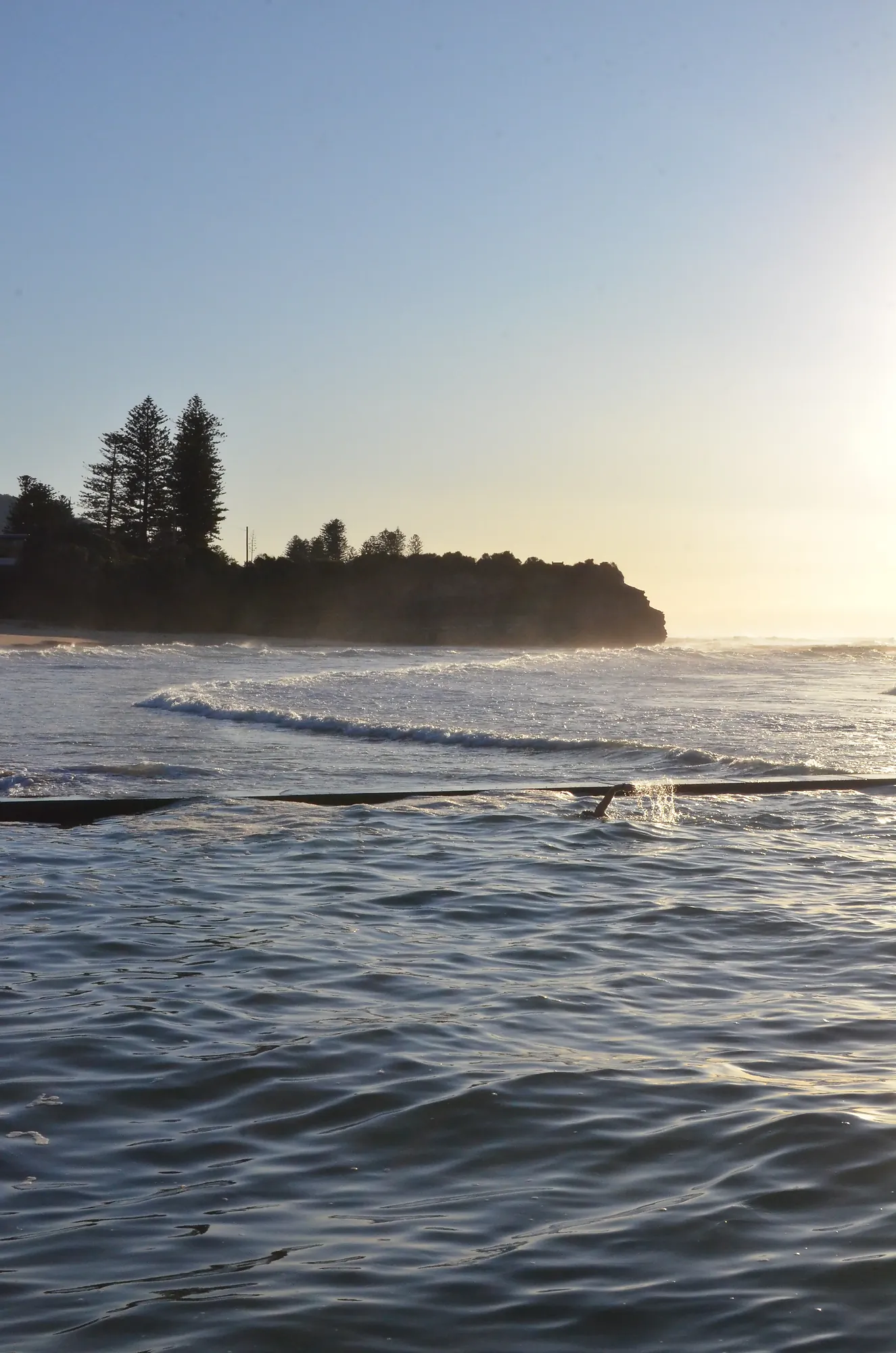 Ocean pool with a swimmer splashing water near a sunlit rocky coastline and trees.