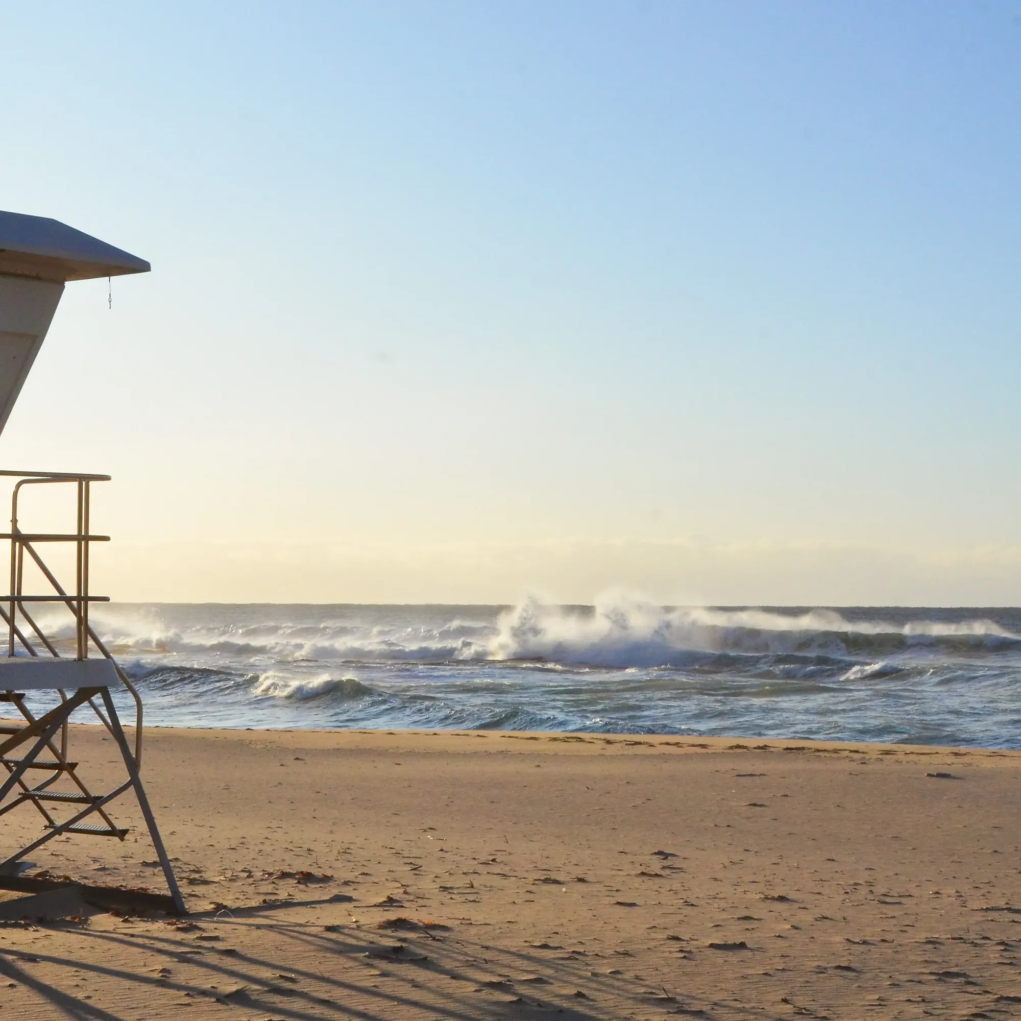 Empty sandy beach with a lifeguard tower on the left and waves crashing in the ocean under a clear sky.
