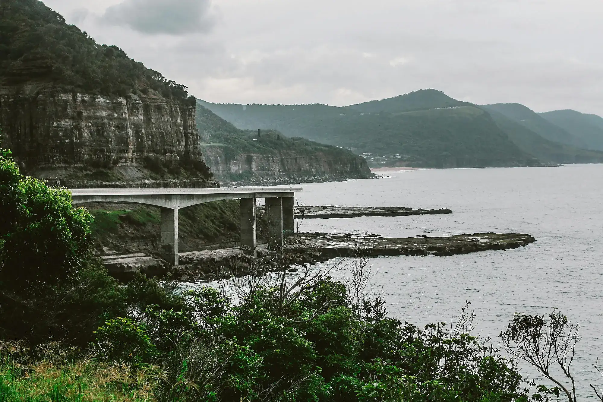 Coastal cliffside with a concrete bridge extending over rocky shore and ocean under cloudy sky.