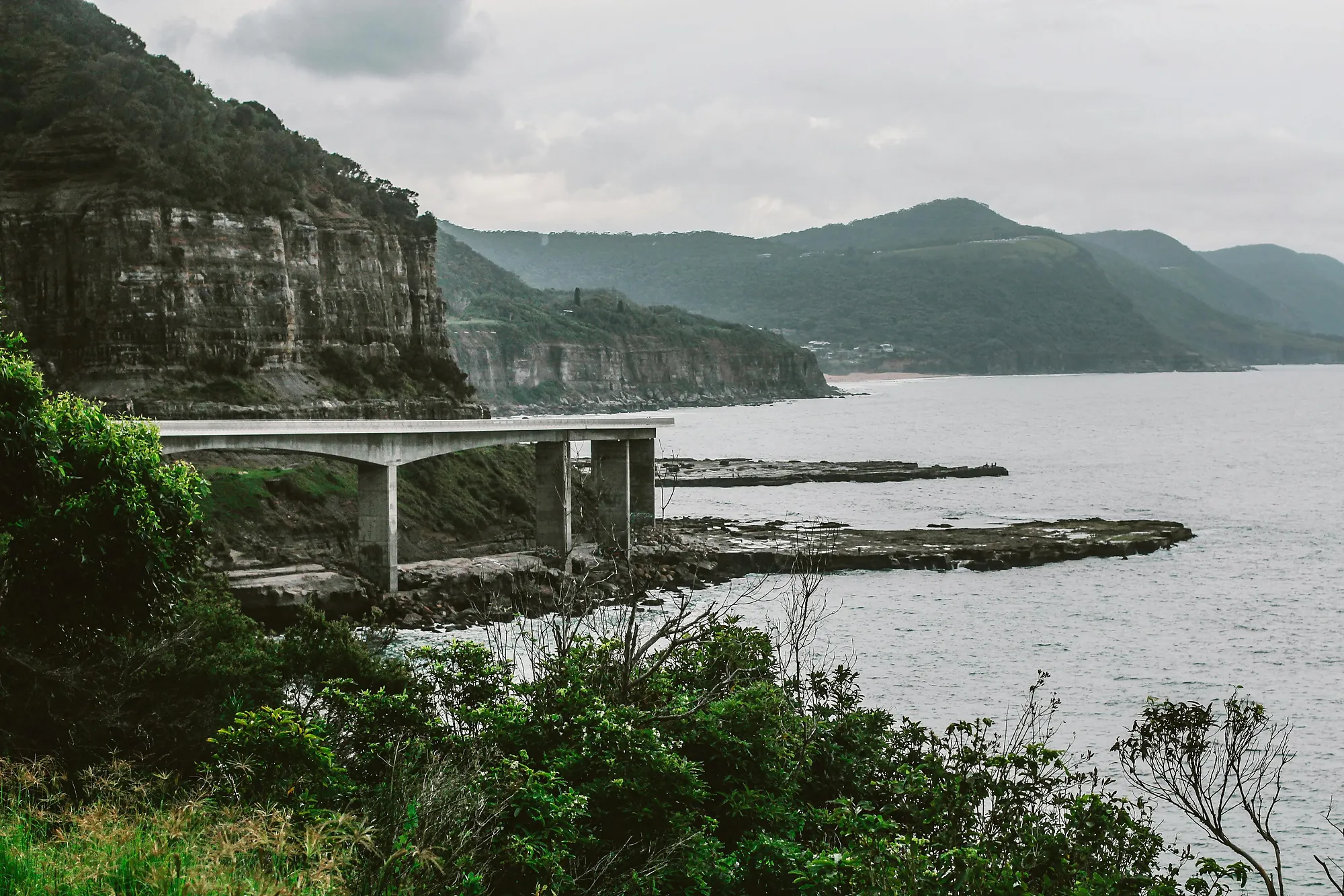 Coastal cliffside with a concrete bridge extending over rocky shore and ocean under cloudy sky.