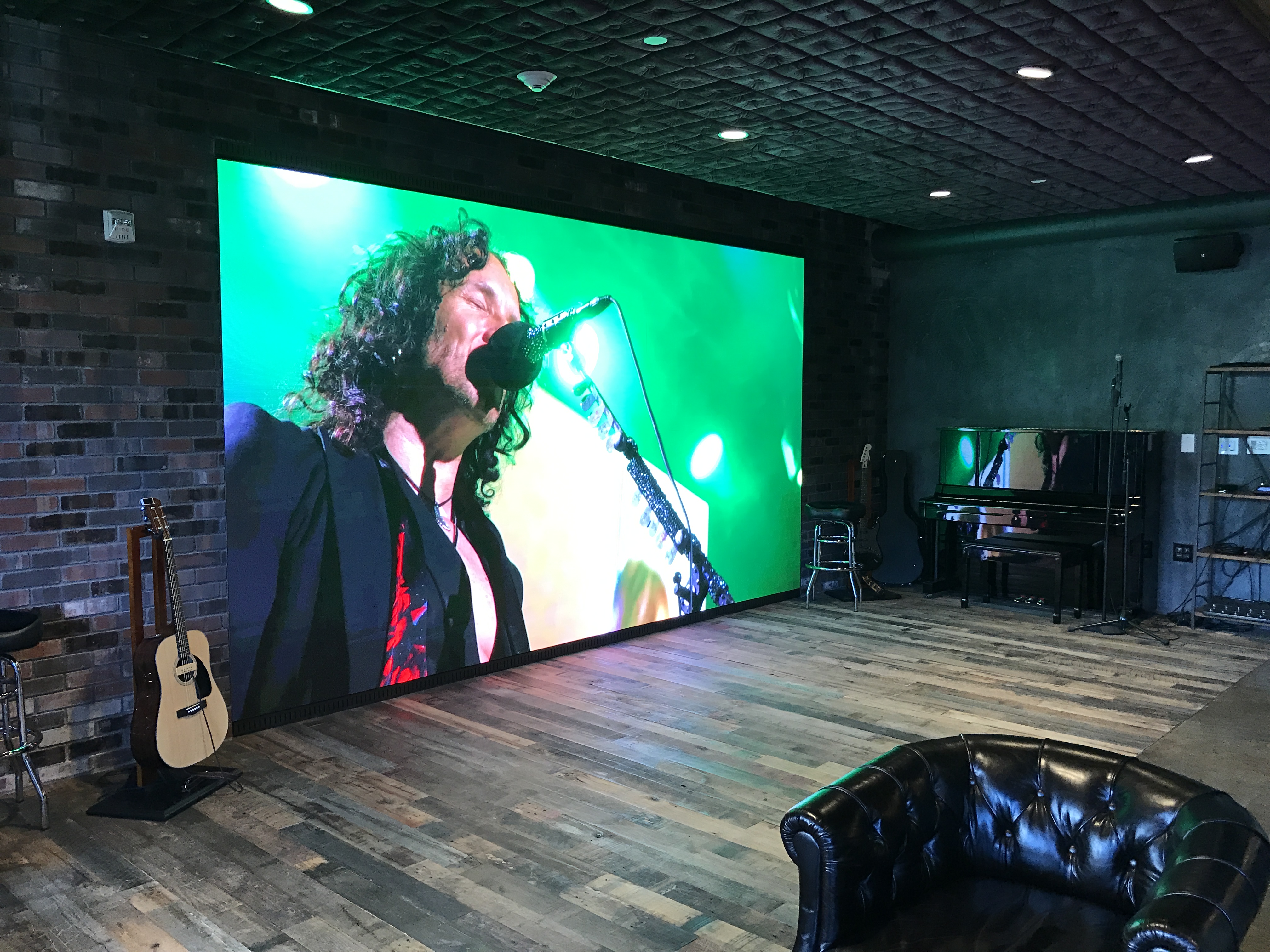Modern room with a large screen showing a musician singing into a microphone, acoustic and electric guitars nearby, a piano, and a black leather armchair.