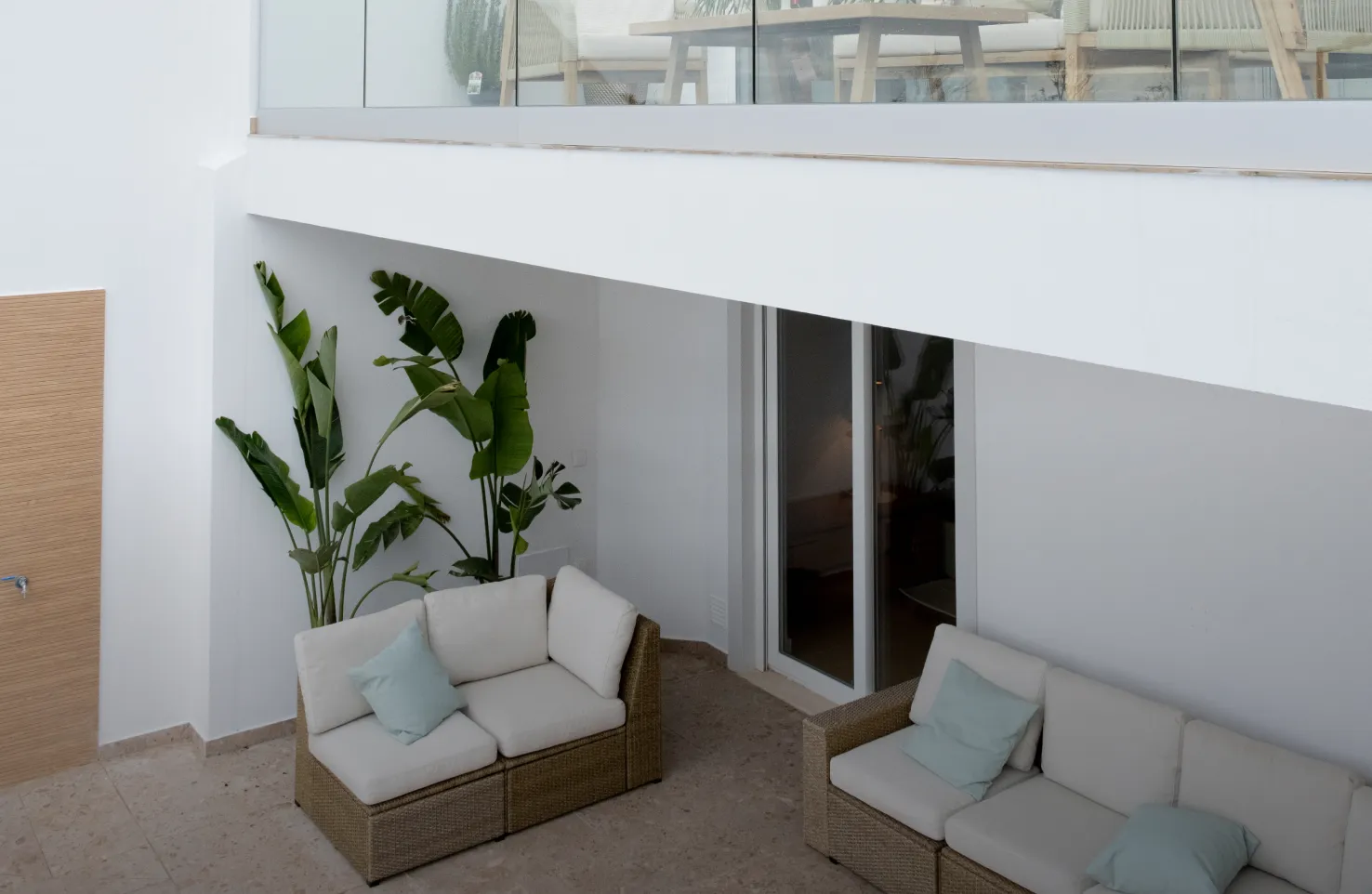 Minimalist white outdoor patio with sofas, plants, and glass railing on upper terrace.