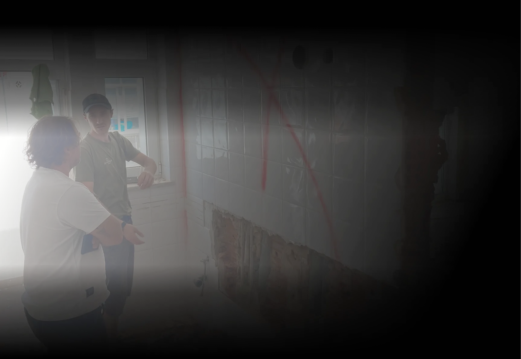 Two construction workers discussing renovation plans inside a tiled kitchen wall under demolition.