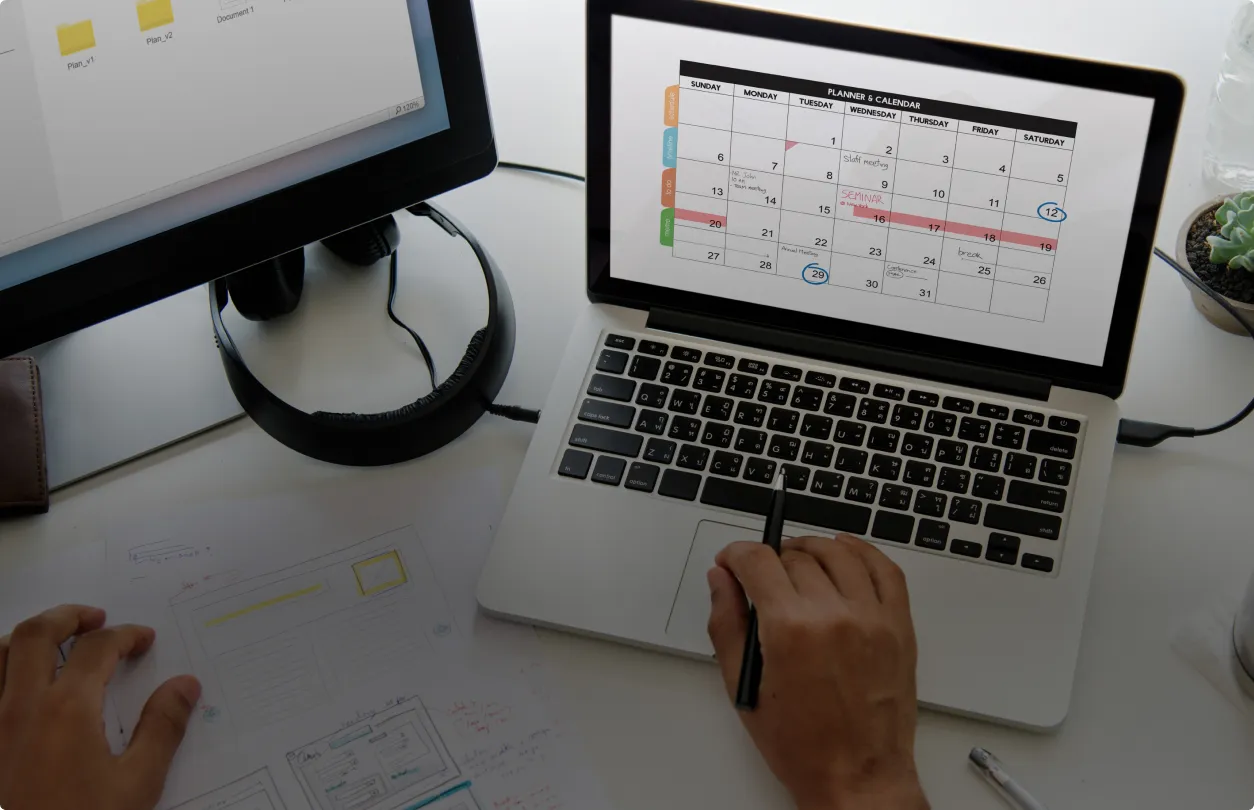 Image of a laptop and desktop on a desk with a calendar on screen and project sketches on paper.