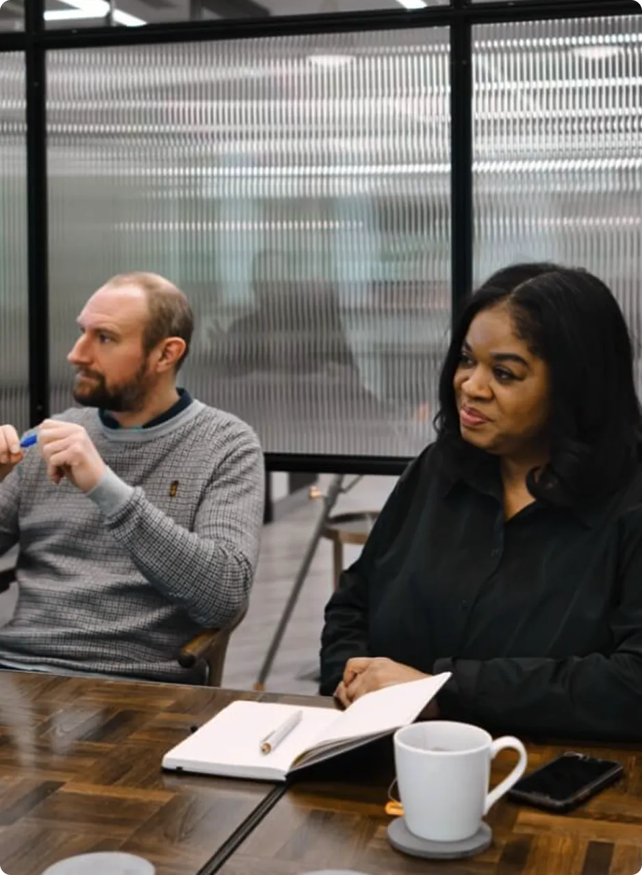 Two professionals sitting at a wooden table in a modern office, engaged in a discussion with a notebook, pen, and coffee cup in front of them.